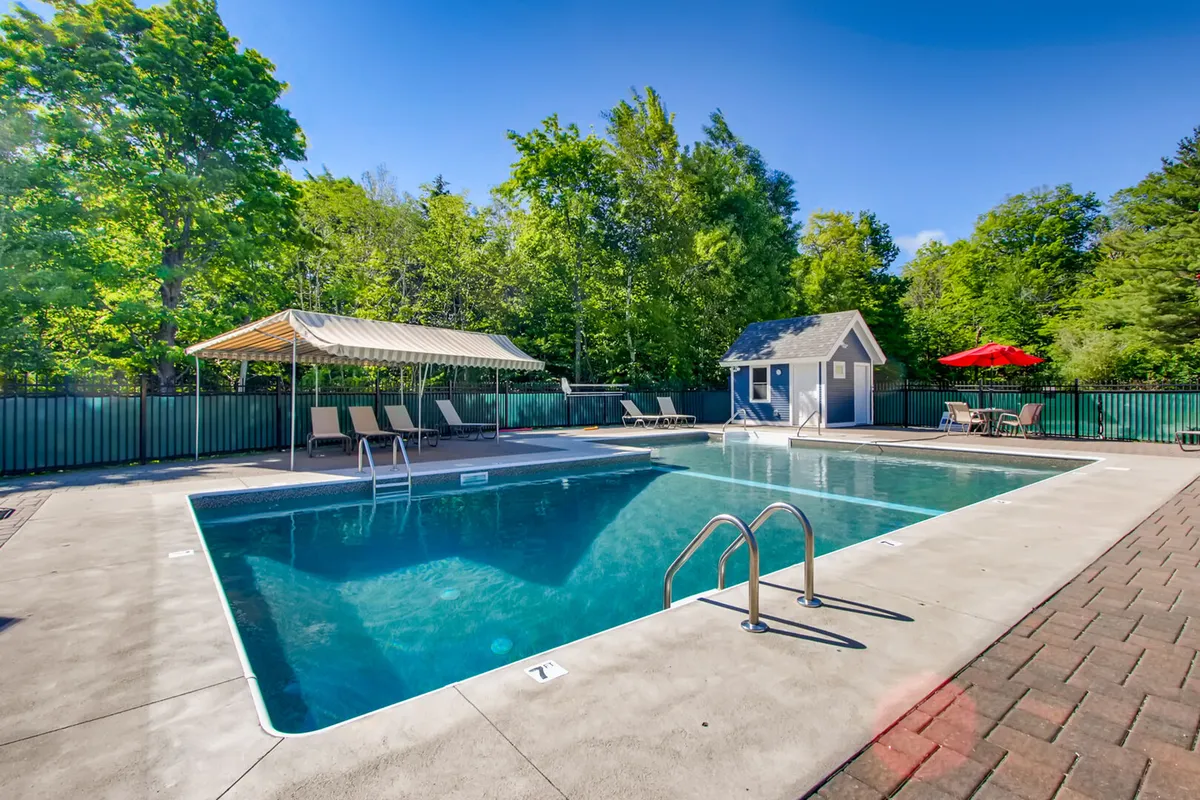 A leisurely outdoor pool scene featuring a sparkling swimming pool, lounge chairs, a canopy-covered seating area, a small shed, and a red umbrella, surrounded by greenery. The atmosphere is relaxed and inviting, perfect for both quiet afternoons and social gatherings.