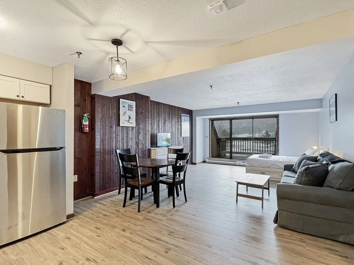 The interior shot features a cozy dining table beside a comfortable sofa, all set in front of a large window that fills the space with natural light. The kitchen area is conveniently nearby, making it easy to prepare meals. The inviting setup suggests the perfect spot for relaxing, sharing meals, reading, or enjoying the view after a day on the slopes.