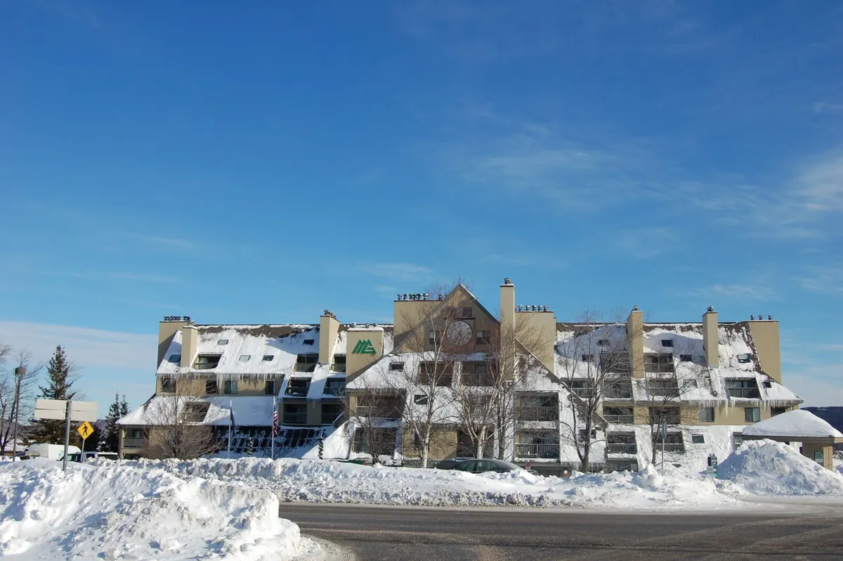 A cozy, snow-dusted building with a distinctive architectural style sits beneath a crisp blue sky. The scene suggests a welcoming lodge atmosphere, with the suggestion of après-ski relaxation awaiting inside after a day on the mountain.