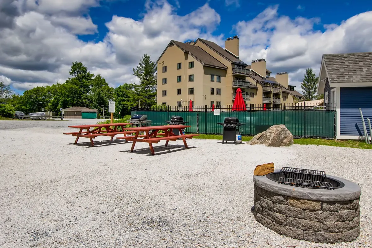An inviting outdoor area featuring picnic tables, a fire pit, grilling stations, and a building in the background. The setting is designed for communal barbecues, evening gatherings, and summer relaxation.