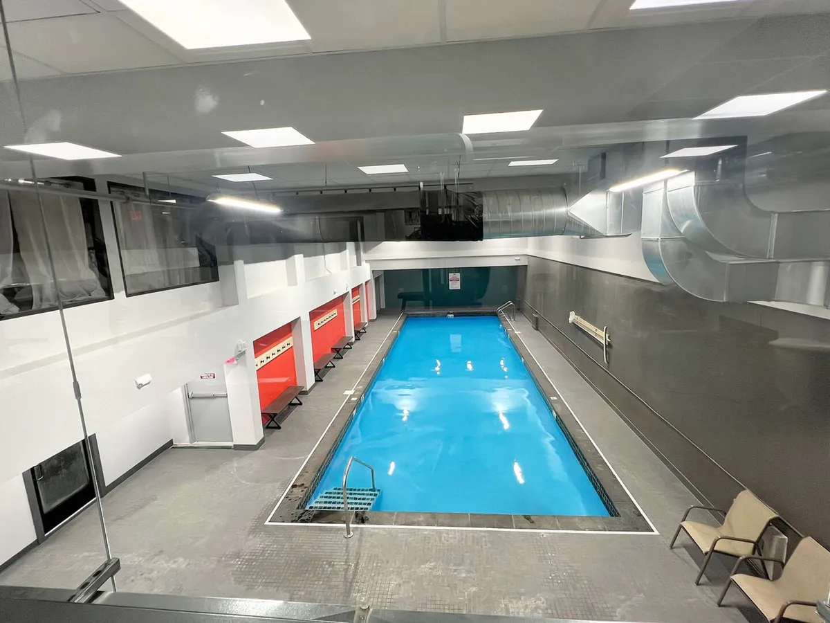 A bright and spacious indoor swimming pool with sparkling blue water, surrounded by red and white changing rooms and empty chairs set along the pool deck. The area is well-lit, exuding a clean and inviting atmosphere ideal for swimming and relaxation throughout the year.