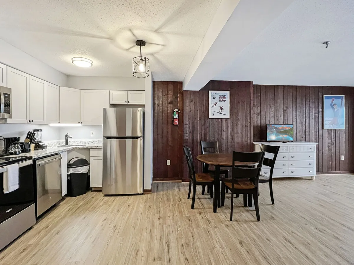 A cozy, modern kitchen area featuring contemporary appliances, a dining table set for meals, and a warm wood panel wall. Decorative posters provide a welcoming touch, while the layout clearly integrates the kitchen with the living and dining spaces, encouraging social interaction and convenient meal preparation.